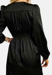 Sieviete melnā garu piedurkņu kleitā ar mazām baltām punktiņām, sasietu vidukli un pufīgām pleciem, roka atbalstīta uz gurniem.