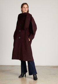 Bordeaux wollen jas met een hoge kraag, lange mouwen, vier grote knopen, zijzakken en een oversized sjaal over de schouders gedrapeerd.
