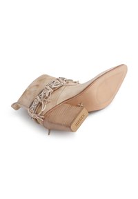 Beige semsket ankelstøvel med stablet hæl, spiss såle, dekorativt sølvspenne og flettet snøredetalj rundt øvre del.