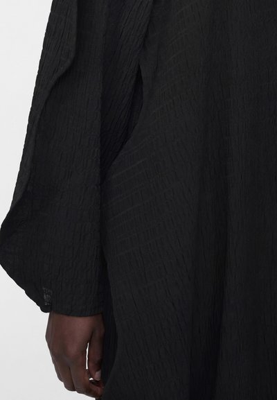 Χέρι με σκουρόχρωμο δέρμα, εν μέρει ορατό κάτω από ένα χαλαρό, υφασμάτινο μαύρο μακρυμάνικο ρούχο σε ανοιχτό φόντο.