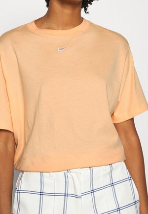 Personne portant un t-shirt Nike à manches courtes couleur pêche et un pantalon blanc taille haute avec des lignes en quadrillage bleu.