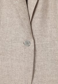 Blazer beige texturizado con un solo botón gris claro abrochado en la parte frontal central, mostrando solapas y dos bolsillos de abertura.