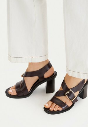 Schwarze Leder-Sandalen mit Absatz, breiten Riemen und goldener Schnalle, getragen zu weißen Hosen, die nackte Füße mit hellem Nagellack zeigen.