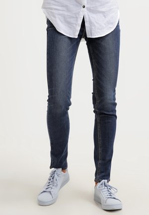 Persoon die donkere skinny jeans draagt, gecombineerd met witte sneakers en een wit overhemd met knopen, staand op een effen lichte achtergrond.