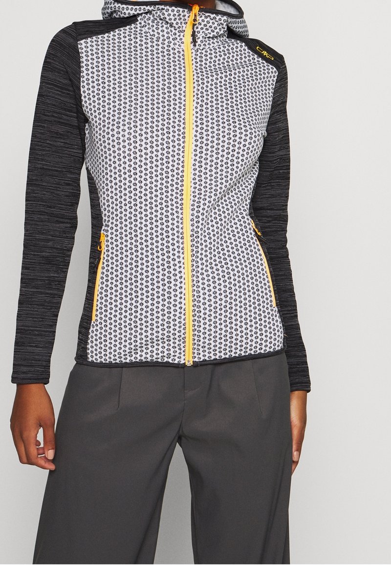 Vrouw in een zwart-wit gestreepte zip-up jack met gele rits en zakaccenten, gecombineerd met donkergrijze broek.