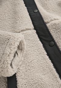 Crème-kleurige fleece jack met zwarte knoopdetails en een gladde stofpaneel aan de voorkant voor contrast. Gelaagde stof biedt extra warmte.