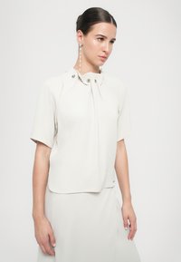Blusa crema a maniche corte con colletto alto, caratterizzata da dettagli drappeggiati e bottoni a pressione argentati. Tessuto morbido con design elegante.