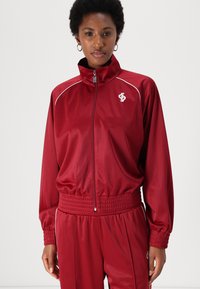 Red track jacket with high collar, zip closure, elastic cuffs, and waistband. Features white piping and logo on the left chest.