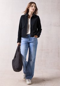 Veste zippée noire avec deux poches avant, associée à un jean large bleu clair et des baskets blanches. Le modèle porte un sac fourre-tout gris foncé.