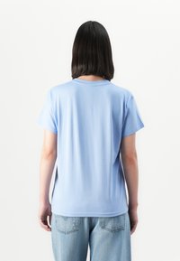 Person with straight black hair wearing a light blue short-sleeve shirt and light blue jeans, standing facing away on a white background.