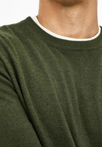 Olivgrüner Strickpullover mit einem gerippten Rundhalsausschnitt und strukturierter Stoffoberfläche. Die Ärmel sind leicht umgeschlagen und geben einen weißen Innenbesatz frei.