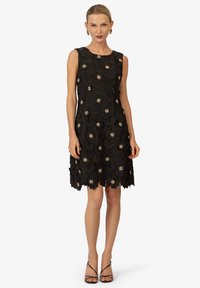 Schwarzes ärmelloses Kleid mit floralen Spitze-Design und bestickten goldenen Akzenten, ausgestattet mit einem gewellten Saum und einer figurbetonten Silhouette.
