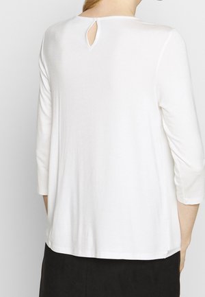 Kobieta ubrana w luźną białą bluzkę z długim rękawem z wycięciem w kształcie łezki na górnej części pleców oraz czarne spodnie.