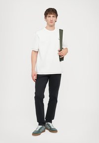 White short-sleeve t-shirt with contrasting stitch details, paired with dark navy pants and green suede sneakers with white laces.