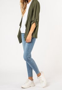 Femme portant une veste olive retroussée, une chemise blanche, un jean skinny bleu clair et des baskets blanches, se tenant sur une jambe.