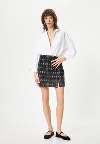 Chemise blanche à boutons avec les manches retroussées, assortie d'une mini jupe à carreaux noir et blanc avec une fente sur le côté. Des ballerines noires complètent la tenue.