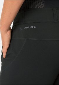 Pantalones de senderismo negros con una textura suave, costuras curvadas y una cinturilla con el logo de Vaude en gris. Incluye una mano en un bolsillo lateral.