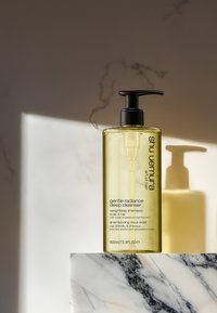 Flacon pompe transparent contenant un nettoyant jaune, étiqueté "nettoyant profond douceur radieuse." Pose sur une surface en marbre, projetant des ombres douces.