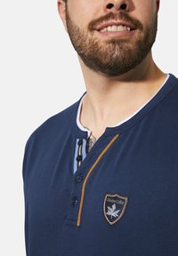 Navy blue cotton henley shirt featuring a logo patch, button closure, and contrasting collar detailing in blue and brown.