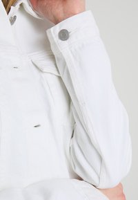 Personne portant une veste en denim blanche avec un bouton-pression argenté au poignet, le bras plié, reposant sur les genoux contre un fond gris.