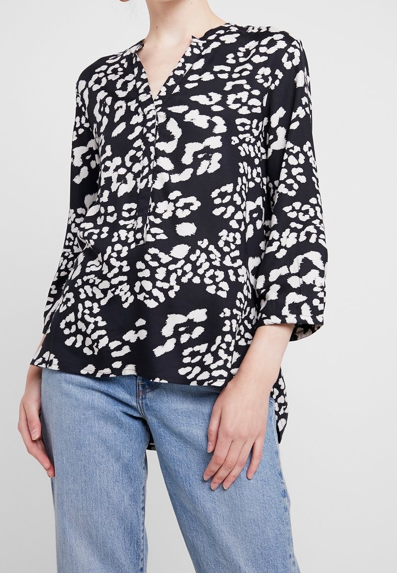 Nainen, joka pukeutuu musta-valkoiseen abstraktiin kuosiset paitaan, jossa on kolme neljännestä hihoista ja vaaleansiniset farkut, seisoo tasaisella taustalla.