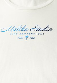 T-shirt en coton blanc avec texte imprimé bleu : "Malibu Studio," "TROUVER LA CONTENTEMENT," accompagné d'un graphique de palmier. Design simple et décontracté.