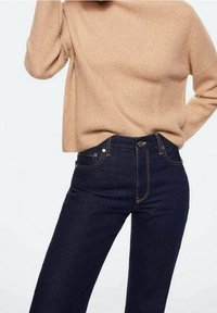 Pull en maille beige associé à un jean bleu foncé taille haute avec coutures orange et poches avant.