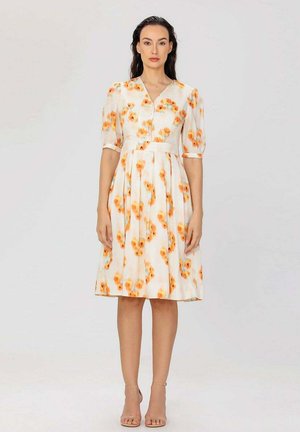 Femme debout, portant une robe blanche mi-longue avec des motifs floraux orange, des manches bouffantes et des détails de boutons sur un fond clair uni.