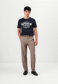 Marineblauwe t-shirt met witte print, gecombineerd met beige broek. De broek is op maat gemaakt, heeft een gladde textuur en een eenvoudig ontwerp. Zwarte schoenen maken de look compleet.