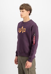 Ljubičasti dukserica s prednjim grafičkim logotipom koji glasi "INDUSTRIES", rebrasti manžeti i rukav s patentnim zatvaračem i crvenom oznakom.