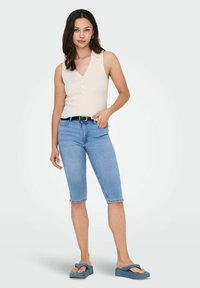 Top senza maniche di colore beige chiaro, a coste, con colletto e bottoni, abbinato a pantaloni corti di denim blu chiaro fino al ginocchio e sandali color teal.