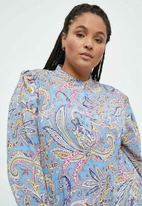 Lyseblå bluse med mønster, som har indviklede paisley-designs i pink, gul og marineblå, med en ståkrave og fine plisseringer på ærmerne.