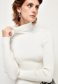 Bia�ły sweter z golfem i długimi rękawami w prążki. Gładka faktura i dopasowany fason. Modelka trzyma kołnierz, pokazując subtelną elastyczność.