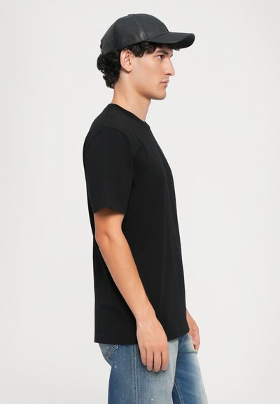 T-shirt nera in cotone a girocollo con maniche corte, abbinata a jeans di denim blu chiaro. Il modello indossa un cappellino nero, visto di lato.