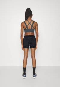 Grå sportbh med korslagda axelremmar, kombinerad med svarta shorts. Modellen står med ryggen vänd mot betraktaren, iförd svarta strumpor och sneakers.