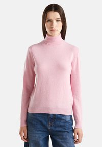Femme portant un pull col roulé rose clair et un jean bleu, se tenant devant un fond blanc uni.