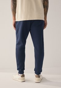 Tengerészkék melegítőnadrág bordázott mandzsettákkal, pamut keverék anyagból, oldalzsebekkel. Krémszínű sportcipőkkel párosítva, barna részletekkel.