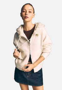 Jasno kremowa bluza na zamek błyskawiczny z puszystym białym kołnierzem, ściągaczami na mankietach i sznurkami. Noszona na ciemno brązowej bluzce i dżinsowej spódnicy.