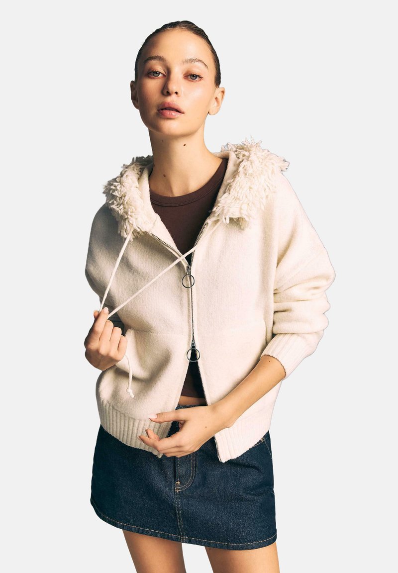 Jasno kremowa bluza na zamek błyskawiczny z puszystym białym kołnierzem, ściągaczami na mankietach i sznurkami. Noszona na ciemno brązowej bluzce i dżinsowej spódnicy.