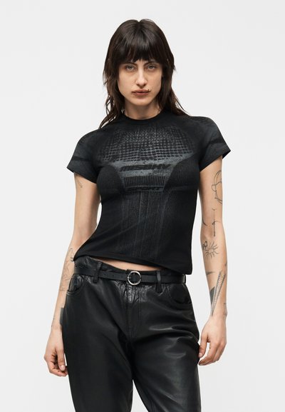 Woman with dark hair wearing a fitted black patterned short-sleeve top and black leather pants with a round buckle belt, standing against a plain background.