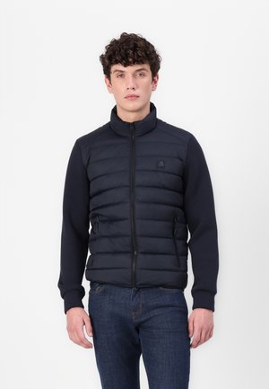 Marineblauer, gesteppten Mantel mit hohem Kragen, Reißverschluss vorne und dunklen Ärmeln; der Stoff wirkt glatt und hat an jeder Seite Taschen.