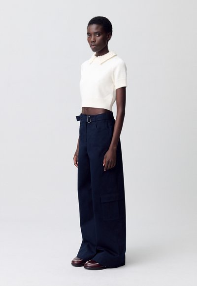 Suéter blanco corto con un cuello texturizado, combinado con pantalones de pierna ancha en azul marino que cuentan con bolsillos laterales y una cintura con cinturón. Zapatos marrones con detalles.