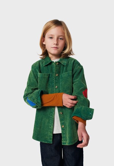 Kind mit schulterlangem blonden Haar, das ein grünes Cordhemd über einem orangefarbenen Langarmshirt und dunklen gestreiften Hosen trägt und den Ärmel anpasst.