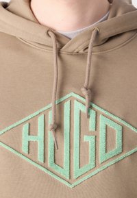 Felpa khaki con cappuccio caratterizzata da un grande logo a diamante verde menta testurizzato con la scritta "HUGO" in lettere in grassetto e con cordini a contrasto.