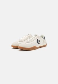 Light beige suede sneakers with a gum rubber sole, featuring a black star logo and white laces. Streamlined design with rounded toe.
