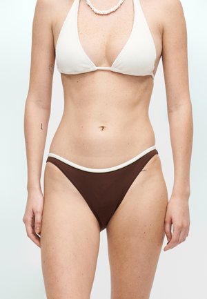 Žena nosiaca biely rebrovaný trojuholníkový horný diel bikin a hnedé spodné diely bikin s bielym lemovaním, stojaca na jednofarebnom svetlom pozadí.