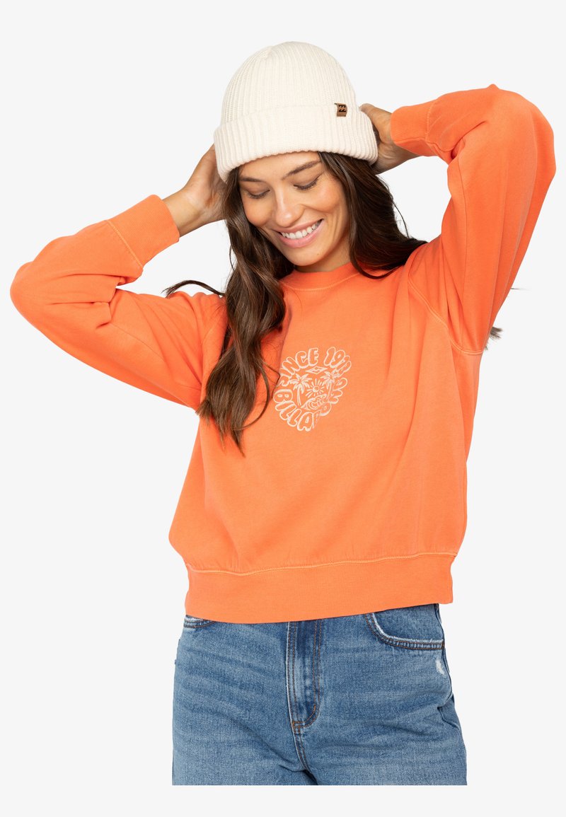 Oranje sweatshirt met geribde boorden en zoom, voorzien van een witte bloemenprint. Gecombineerd met een crèmekleurige gebreide beanie. Blauwe spijkerbroek.
