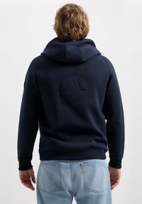 Haut à capuche bleu marine avec un logo circulaire texturé au dos, capuche avec cordon de serrage et poignets côtelés. Fabriqué en tissu doux et durable.