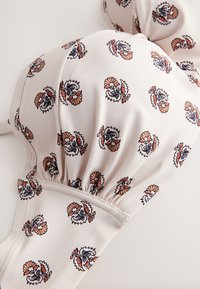 Gros plan d'un haut de bikini blanc avec de petits motifs floraux détaillés en orange, bleu et noir, avec une couture froncée sous les bonnets.