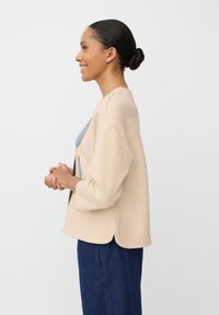 Beżowy sweter z miękką teksturą, okrągłym dekoltem i rękawami do 3/4. Połączony z ciemnymi dżinsami, przedstawiony z boku.